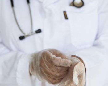 Faceless ethnic medical worker in lab coat and stethoscope taking of transparent gloves after approaching patients for examination in modern hospital